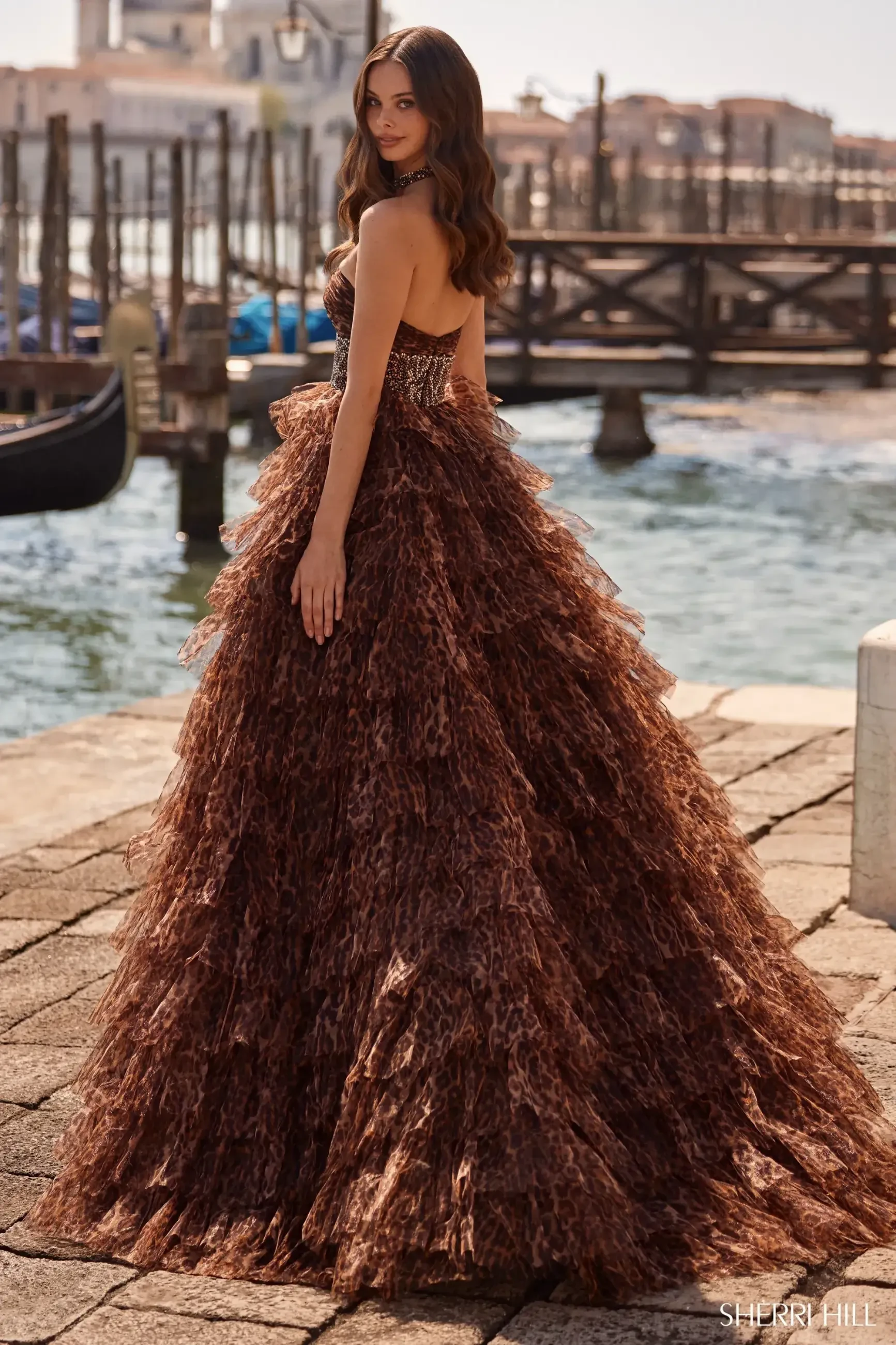 Elegant woman in a layered, textured brown gown poses on a stone walkway before a Venetian bridge. The scene exudes sophistication and charm.